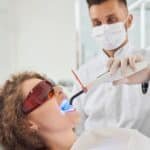 Dentist performing a teeth whitening procedure using a blue LED curing light on a patient wearing protective glasses in a dental clinic.